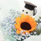 Graduation bouquet with a sunflower, baby's breath, and a graduation cap on a fluffy white decoration.