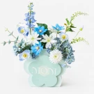 Artificial flower arrangement with blue and white flowers in a flower-shaped box with "MOM" on it.
