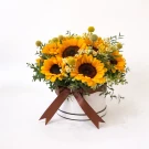 A white circular box filled with bright sunflowers, greenery, and accent flowers, tied with a brown ribbon.