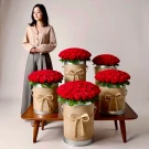Woman posing with five large buckets of red roses on a wooden table.