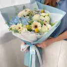 A person holding a Blue Serenity Bliss Bouquet with blue hydrangeas, white roses, and yellow button flowers, wrapped in blue and white paper.