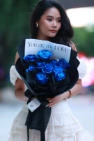 Woman holding a bouquet of vibrant blue roses wrapped in black paper with a card that says "You are my LOVE".