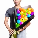 Man holding a bouquet of colorful smiley face balloons wrapped in black paper and tied with a green ribbon.