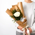 Hand holding a white rose bouquet with eucalyptus, wrapped in brown paper and tied with a white ribbon, against buildings.