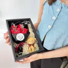 A gift box filled with red soap roses, a teddy bear keychain, and straw.