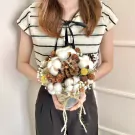 A person holds a rustic bouquet of cotton flowers, pine cones, and yellow accents in a silver pot.