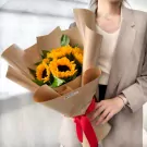 Woman in a blazer holding a bouquet of sunflowers wrapped in brown paper with a red ribbon.