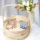 Round woven bamboo basket with lid, filled with food, topped with white flowers and a jasmine garland.