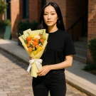 Woman in black shirt holding a bouquet of orange and yellow flowers wrapped in yellow paper with a white ribbon.
