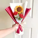 Crocheted bouquet of red roses and a yellow sunflower wrapped in red paper and tied with a cream ribbon.