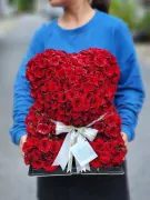 A heart-shaped bear made of fresh red roses, held by a person wearing a blue sweatshirt.