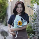 A smiling young woman in glasses holds a graduation bouquet with a sunflower and a small stuffed bear wearing a graduation cap.