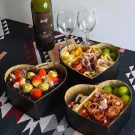 Heart-shaped charcuterie box with grapes, cheese, cured meats, and strawberries, next to wine and glasses.