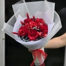 Hand holding a bouquet of deep red roses with white bead accents, wrapped in white paper and tied with a red ribbon.