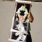 A person holds a gift box containing a large, white jasmine garland with a pink flower centerpiece.