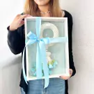 Woman in jeans and black sweater holds a gift box containing a white jasmine malai with blue flowers and ribbon.