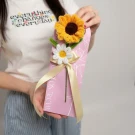 A person holds a pink card with a sunflower and daisy craft, tied with a yellow ribbon.