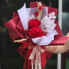 A bouquet of red dried roses and white filler flowers, wrapped in red paper and red mesh with a ribbon.