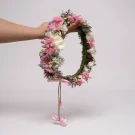 Hand holding a large flower crown with pink and white blooms, featuring a twine ribbon with pink bows.