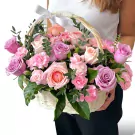 A white woven basket brimming with pink and purple roses, carnations, and greenery, tied with a white ribbon.
