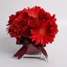 A square glass vase filled with red gerberas, carnations, and roses, tied with a deep red ribbon.