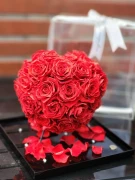 Preserved red roses in heart shape under glass dome, with red petals and pearls on a black tray.