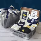 A baby boy's pilot-themed gift set with romper, shoes, bib, and cap inside a silver suitcase.