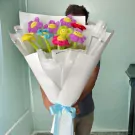 Bouquet of colorful plush smiley-face daisies in white and silver wrapping, tied with a light blue ribbon.