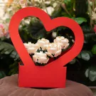 A red heart-shaped basket holding a bouquet of 5 white Lego roses, set against a backdrop of green leaves and pink flowers.
