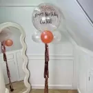 Large clear bubble balloon filled with rose gold glitter, with "Happy Birthday GUJAI" written on it.