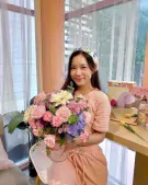 A young woman in peach loungewear holds a pastel flower bouquet with blue hydrangeas, pink roses, and white gerberas.