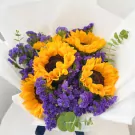 A round bouquet of sunflowers and purple statice flowers wrapped in white paper.