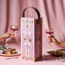 Pink chocolate box with romantic floral and hourglass designs, holding chocolates, with hands holding wine glasses.
