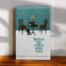 Book cover of "Before the Coffee Gets Cold" by Toshikazu Kawaguchi, featuring a minimalist cafe scene with two chairs, a table, and a cat.