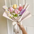 Crocheted bouquet of pink, yellow, and red tulips with white daisies and purple sprigs, wrapped in pale paper and tied with a ribbon.