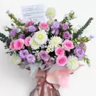 A floral stand with pink roses, purple lisianthus, white dahlias, and green foliage, wrapped in pale pink and white paper.
