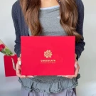 Woman holding a red chocolate box with gold floral design and "CHOCOLATE SAVORY AND SWEET" text.