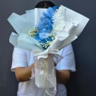 A person holds a dried flower bouquet in shades of blue and white, featuring a rose and delicate baby's breath.