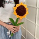 A person holds a large, crocheted sunflower with brown and yellow petals, green stem and leaves.