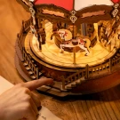 A finger points to a miniature wooden carousel with painted horses and stairs.
