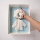 Hand holding white scarf adorned with gold brooch, arranged like a doll in a blue box.