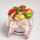 Round white fruit box with apples, kiwi, orange, and pear, tied with a pink bow.