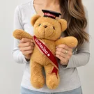 A teddy bear wears a graduation cap and a red sash with Thai writing.