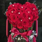 Heart-shaped bouquet of red roses with heart crystals in a clear acrylic cylinder vase.
