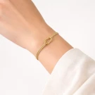 Gold-tone bracelet with an infinity knot on a woman's wrist, paired with a white shirt.
