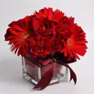 Red gerberas, carnations, and roses in a square glass vase with a burgundy ribbon.