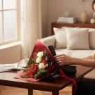 A hand holding a red and black wrapped bouquet of white roses, red gerberas, and pine on a table.