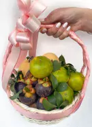 Wicker basket filled with mangosteens, oranges, and pears, adorned with a pink ribbon and held by a hand.