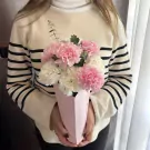 A person in a striped sweater holds a pink and white carnation bouquet in a pink paper vase.