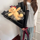 A teddy bear with a graduation hat sits in a bouquet of dried flowers, wrapped in black paper with a burgundy ribbon.